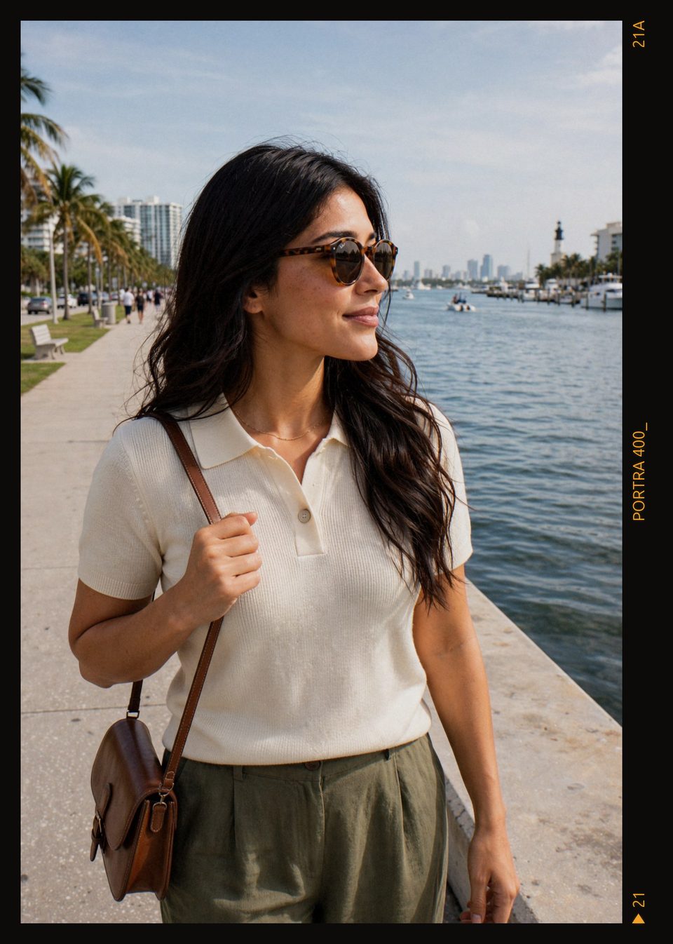 Kaethia_ in tortoiseshell sunglasses and a cream knit polo walks along a waterfront path, looking out to her right toward the water, with palms, boats and city towers behind her.