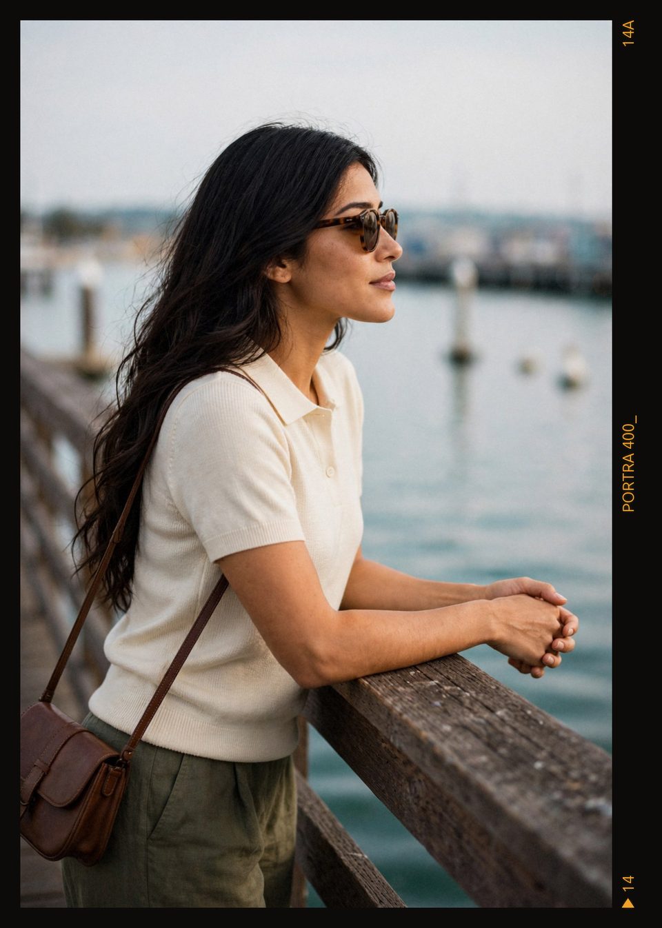Kaethia_ leans on a weathered wooden rail in profile, hands clasped, sunglasses on, gazing out over calm water in soft evening light.