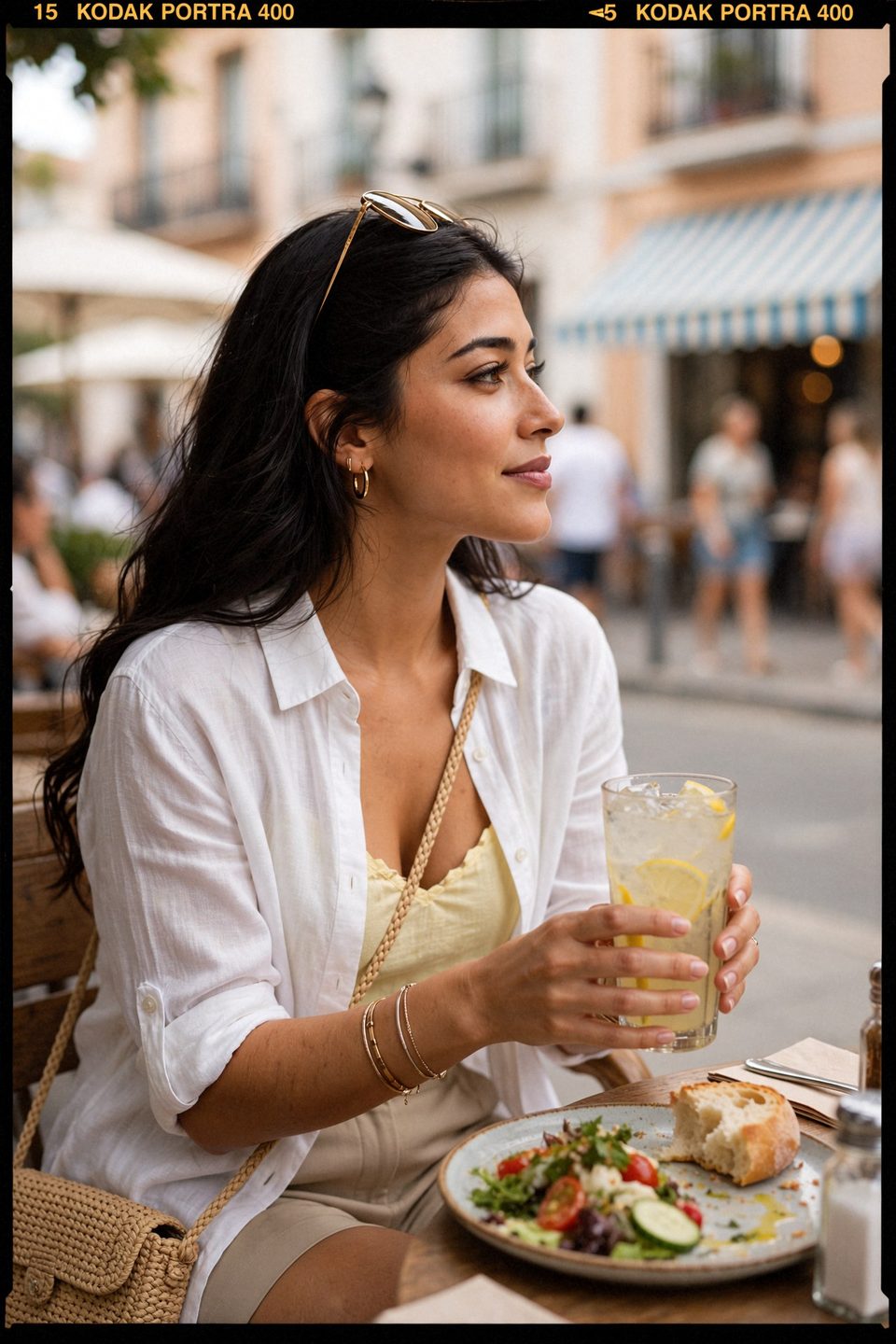 Kaethia_ in a white linen shirt and yellow top sits at an outdoor table in profile, holding a glass of lemonade and gazing up the street, with a salad and bread in front of her and blurred passers-by behind.