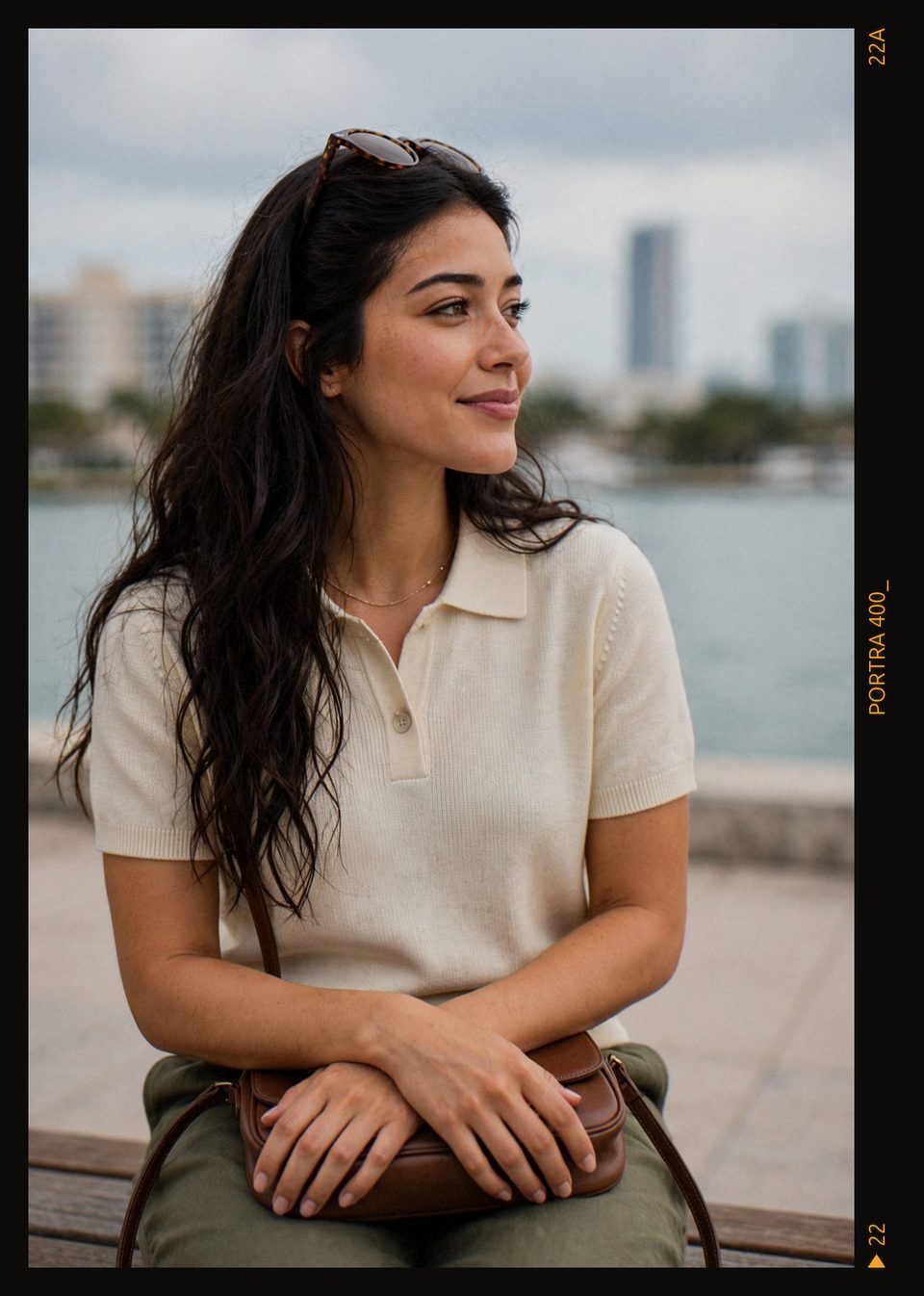 Kaethia_ sits on a bench with her hands resting on a small brown bag, sunglasses pushed into her hair, smiling and looking out toward the water under a cloudy sky.